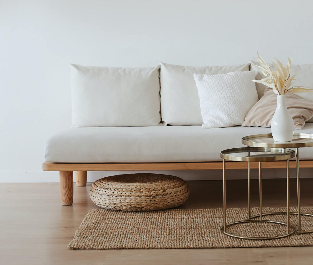 Minimalistische inrichting, een lichte bank op houten pootjes tegen een lege muur met rustige elementen en aankleding. Rond jute kussen, jute vloerkleed en een set ronde gouden salontafels.